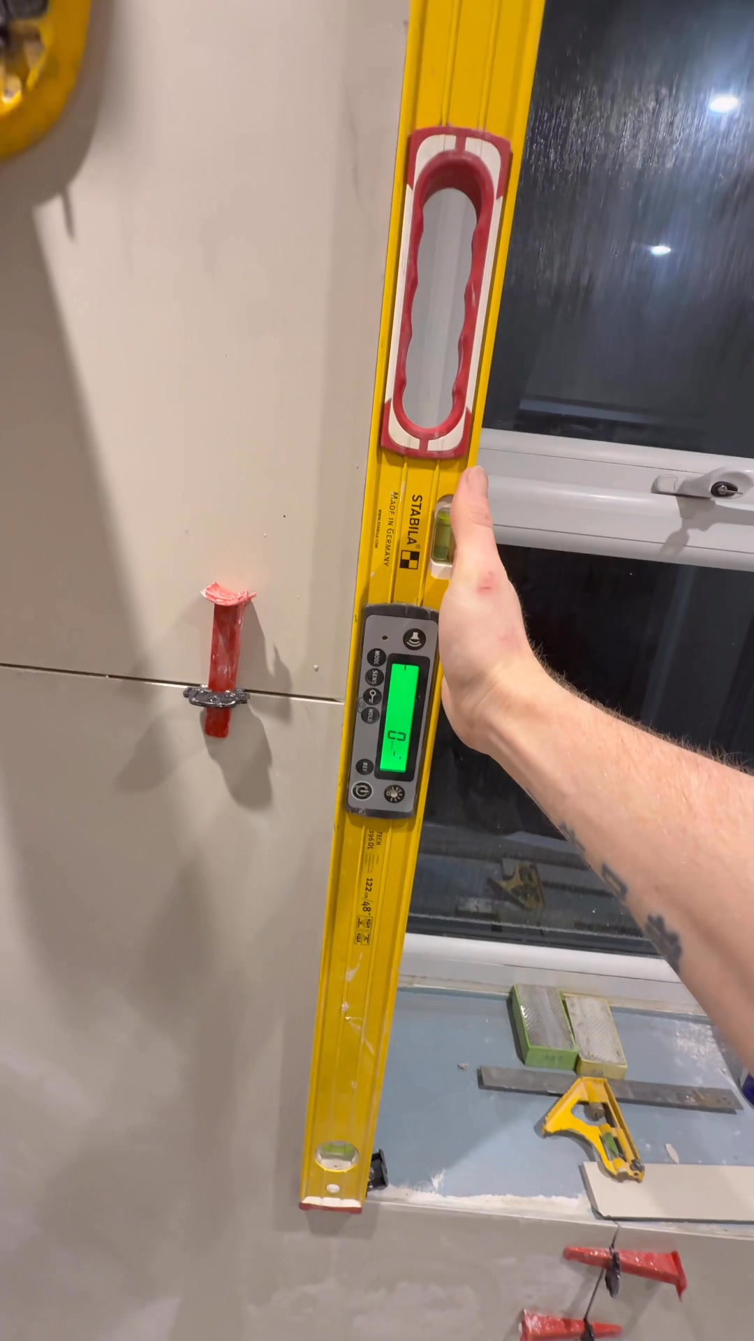 A digital spirit level checks the alignment of large wall tiles beside a bathroom window during installation.