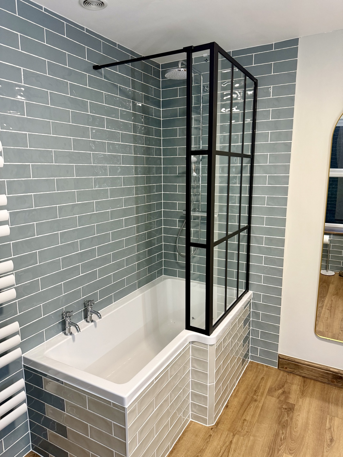 Blue-grey metro tiled bathroom with an L-shaped bath, black-framed glass screen, pale tiled bath surround and wood-effect floor.