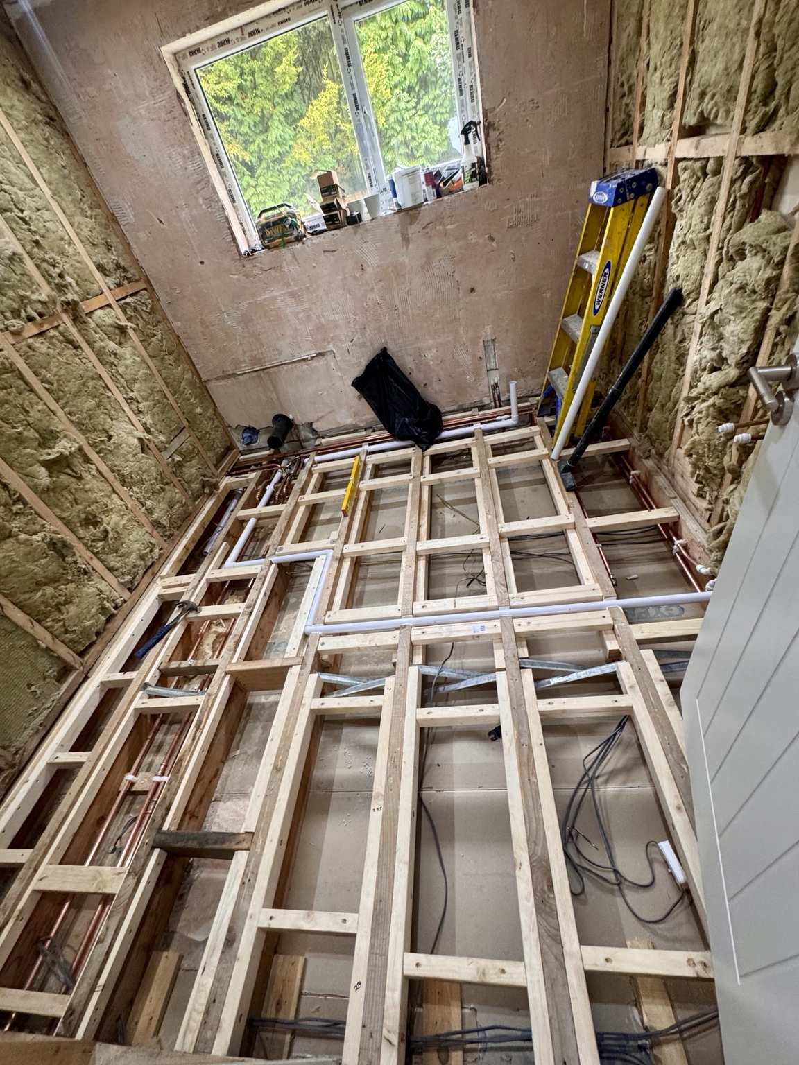 Exposed bathroom floor framing with pipework, cables and insulated wall cavities before the floor is covered.