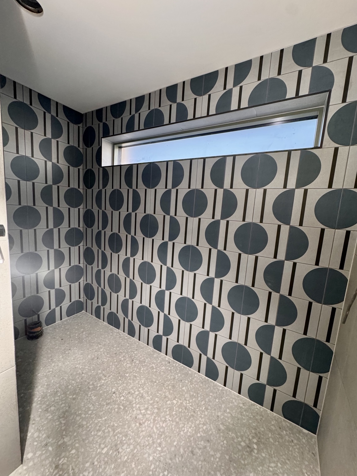 Blue and cream geometric wall tiles around a high window, with a terrazzo-effect floor and an exposed plumbing connection.