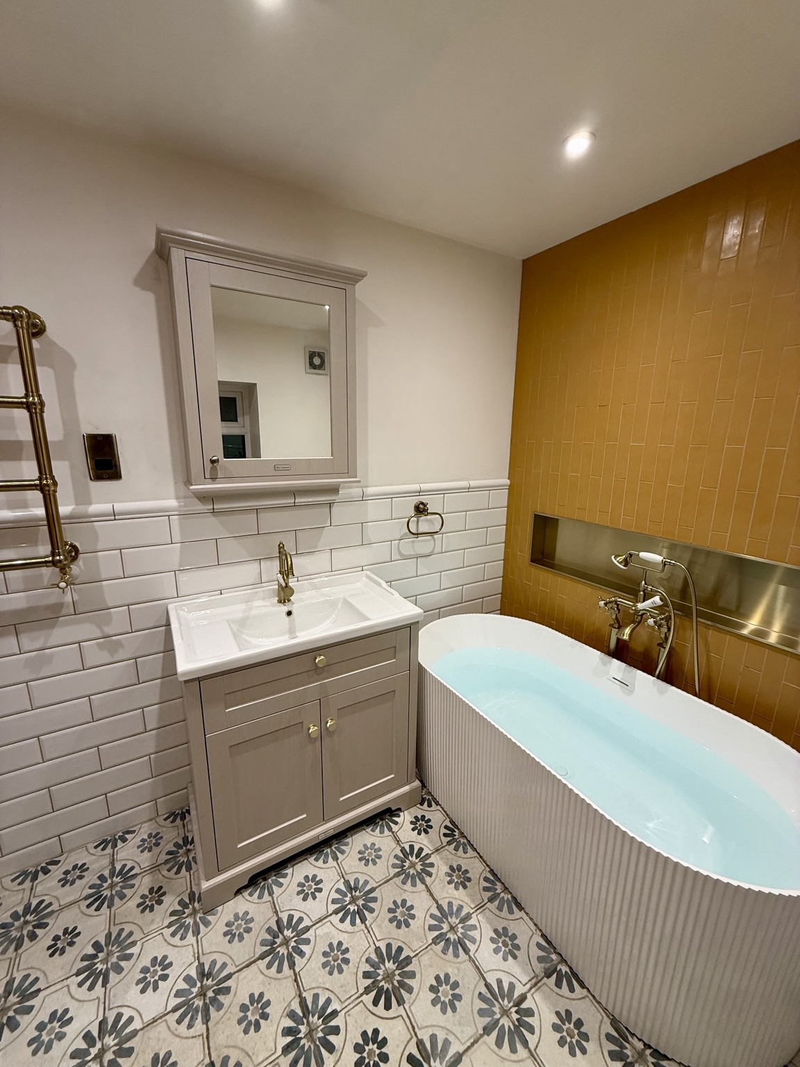 Bathroom with a fluted freestanding bath, mustard feature tiles, brass-coloured fittings, vanity storage and patterned floor tiles.