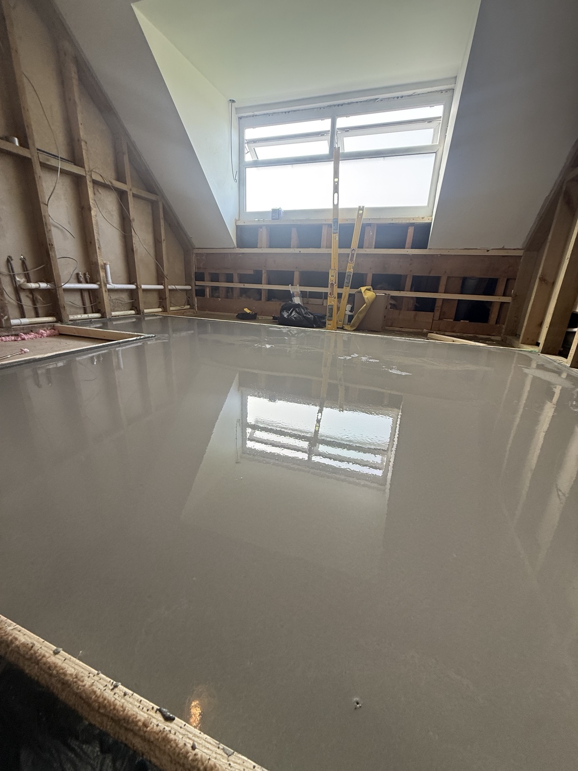 Fresh grey levelling compound across a bathroom floor, with exposed wall framing and pipework beneath a dormer window.