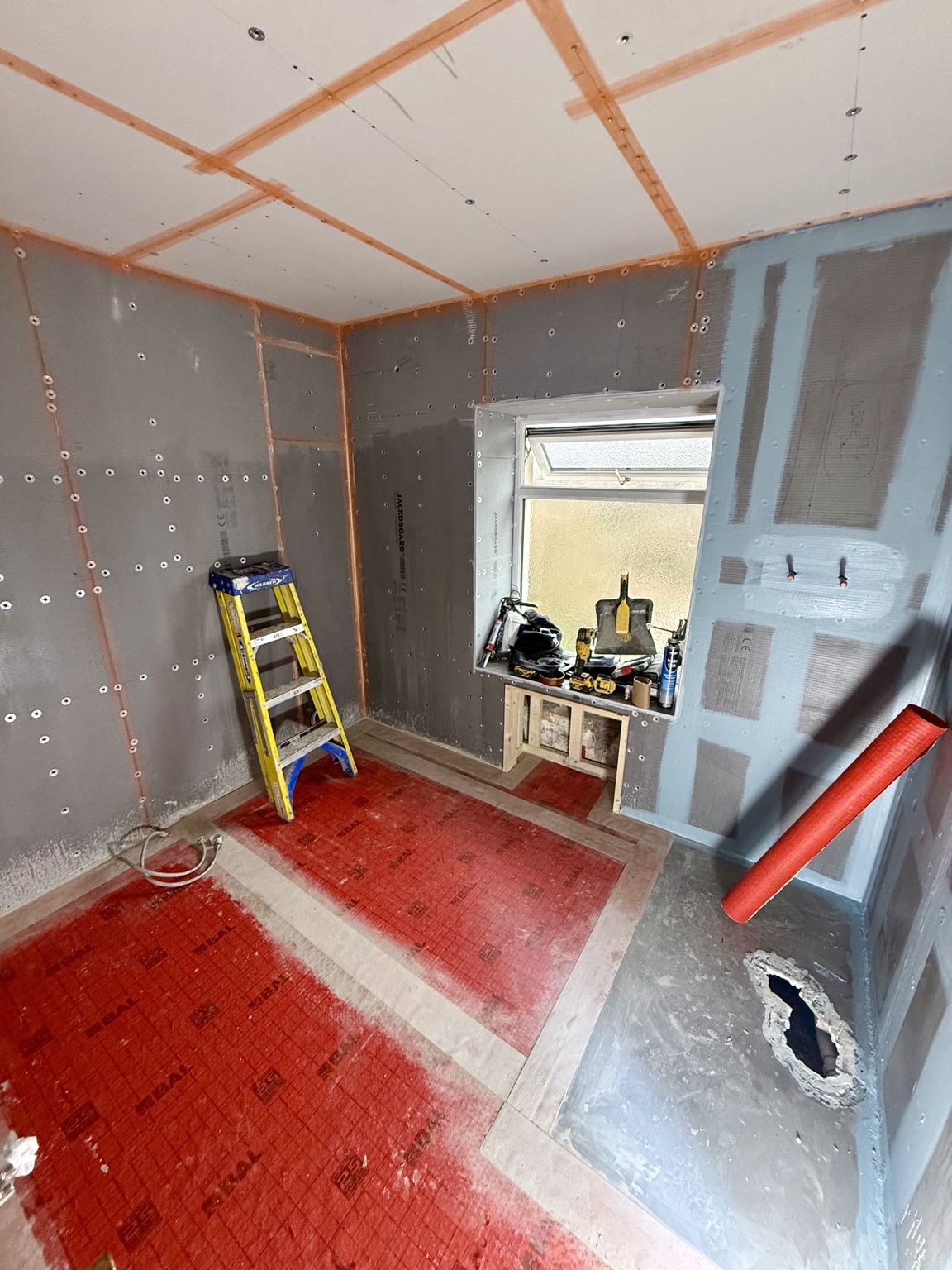Bathroom wall boards with visible fixings and treated joints above a red floor membrane, with tools and unfinished openings visible.