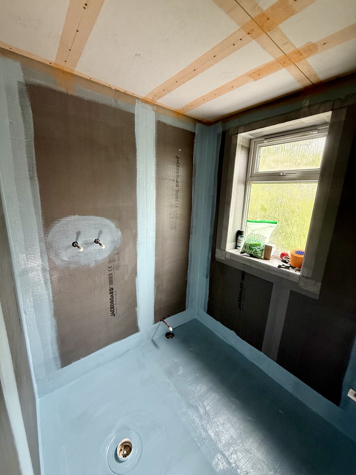 Bathroom preparation showing a blue waterproofing layer on the floor, wall junctions and board seams, with plumbing openings visible.